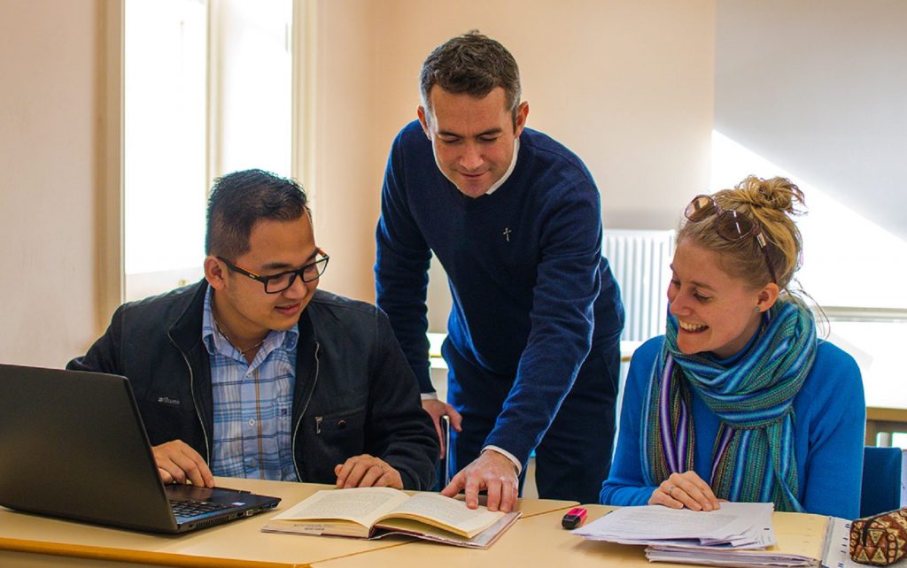Three people talking in a classroom situation. Left: male student sitting down with a laptop in front of him looking at a book to his left. Middle: Rev. Dr Jake Mudge is standing up and leaning down pointing to something in the book. Right: Female student is sitting down with a pile of note papers/books in front of her looking at the open book to her right