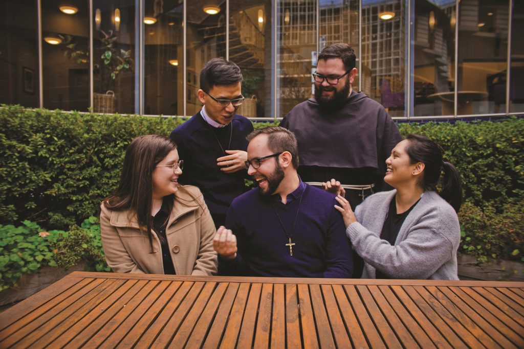 Five students laughing at a picnic table in garden of Thomas Carr Centre with the curved glass wall behind them