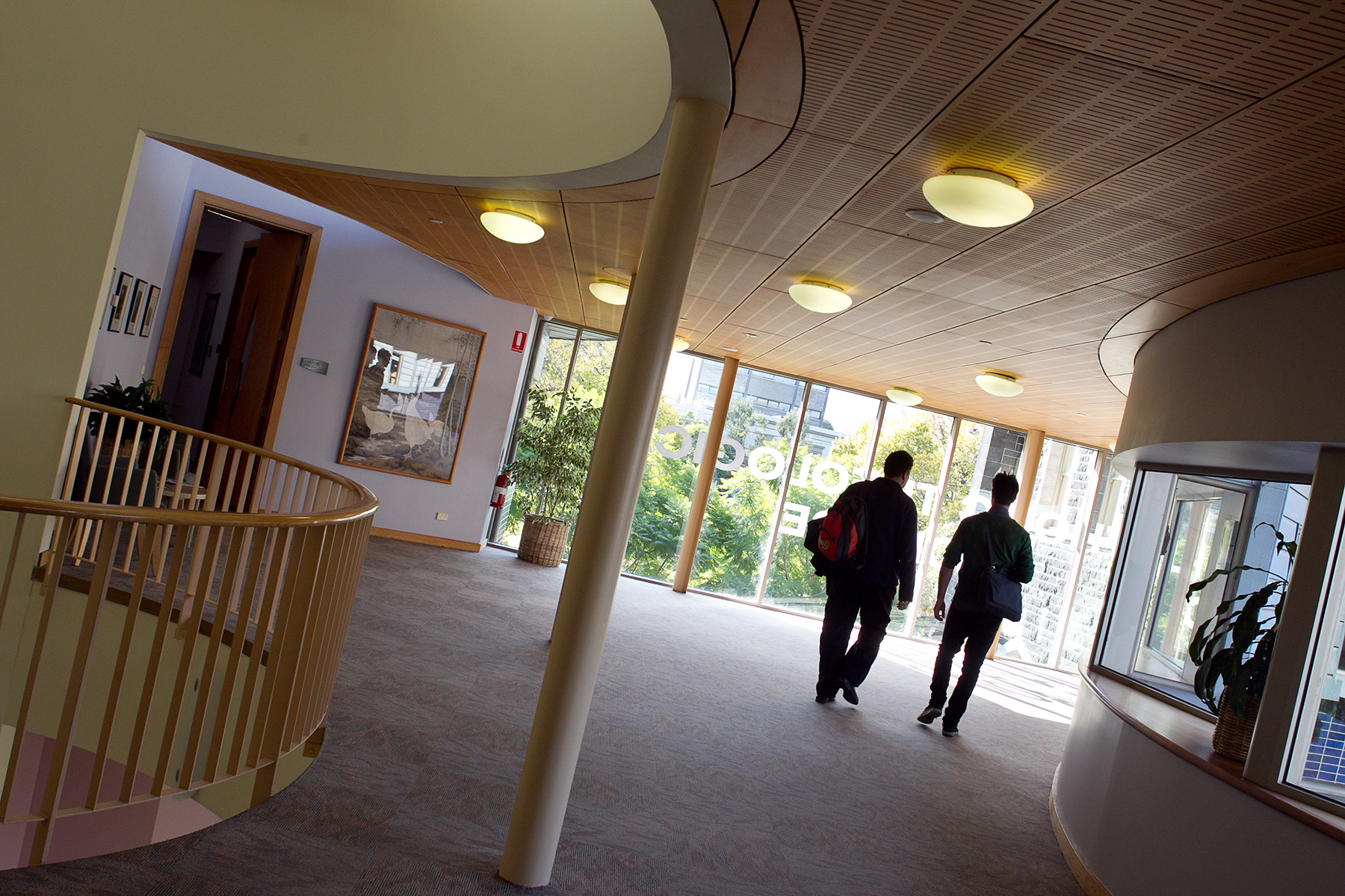 Two male students walking away from the camera in the level 3 foyer towards the wall of windows. Stairs are off to the left