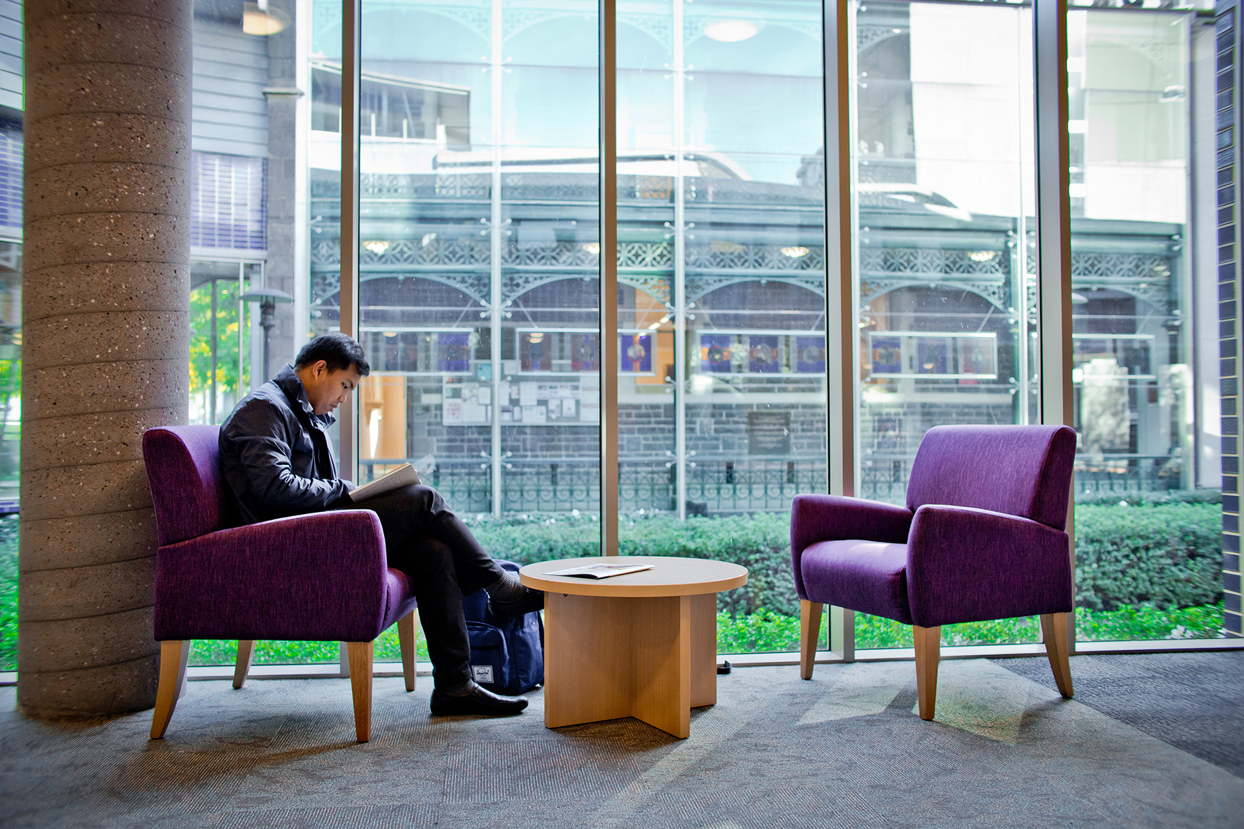 Male student seated on one of two purple chairs by a wooden coffee table in the level 2 foyer by the glass wall/window looking out into the garden