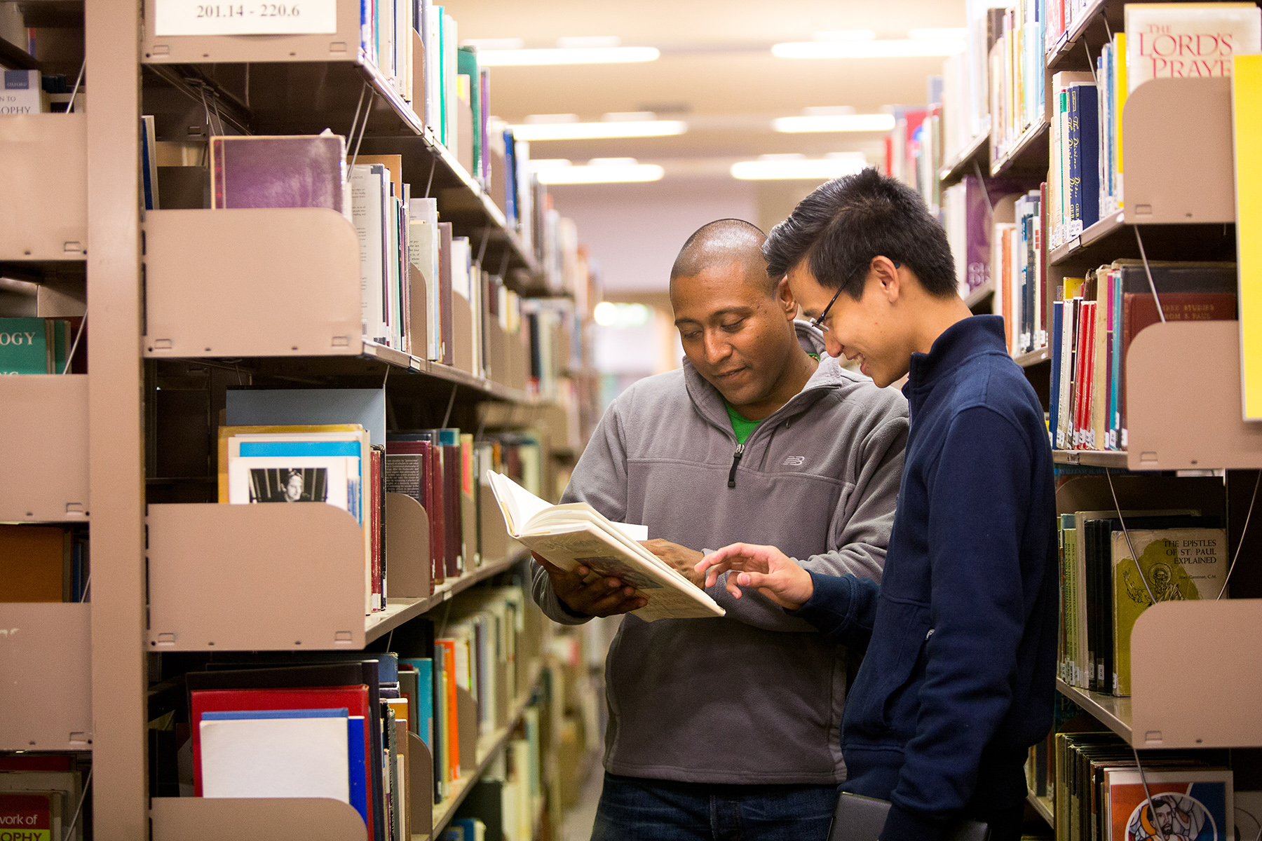 Two male students talking over an open book between shelves of the Mannix Library